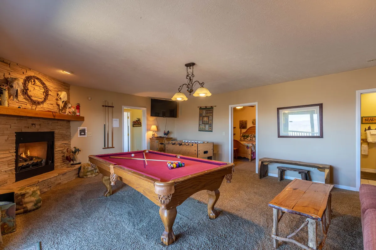 A casual, cozy game area featuring a pool table prominently in the foreground, with a rustic fireplace, wooden bench, and decorative touches enhancing the ambiance. The layout suggests an intimate setting for small-group fun and shared moments.