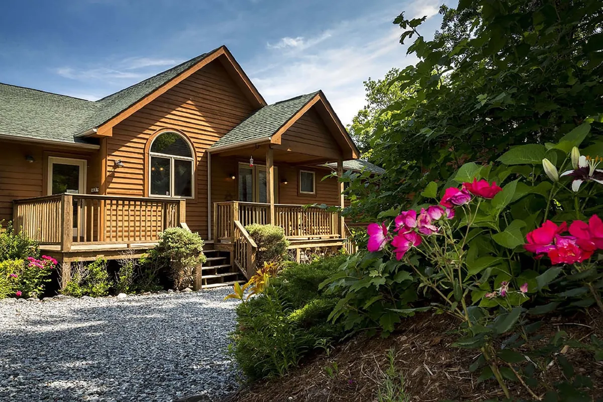 A welcoming wooden porch adorned with vibrant flowers and an arched window. The porch looks out onto a gravel pathway and the lush natural surroundings, with ample space for sitting and socializing outdoors.
