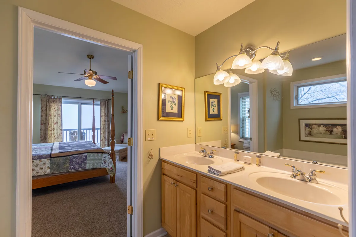 A serene ensuite bathroom with a double sink vanity, decorative mirrors, and framed art. The doorway offers a glimpse of the spacious bedroom beyond, emphasizing privacy and comfort.