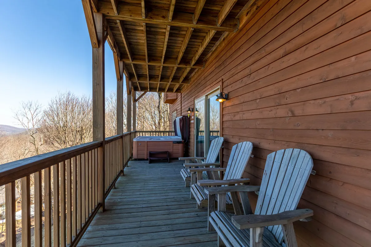A spacious wooden deck set against a backdrop of rolling mountains, featuring inviting rocking chairs and a bubbling hot tub. The deck offers panoramic views for morning coffee or sunset relaxation, and blends seamlessly with the natural woodland surroundings.