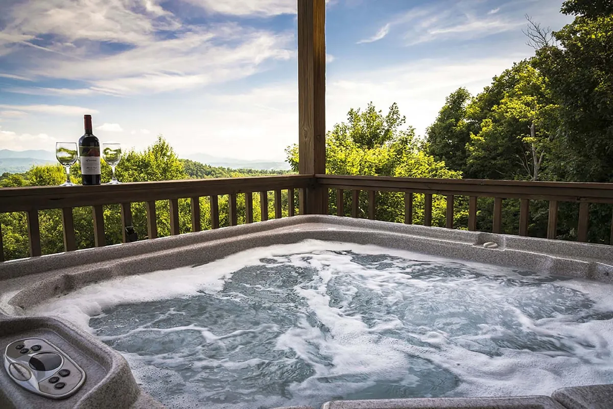 An inviting outdoor hot tub set on a spacious deck, with a pair of wine glasses and a wine bottle nearby. The tub faces out towards a scenic mountain view, capturing the moment at sunset. The setting suggests relaxation and togetherness after a day of exploration.