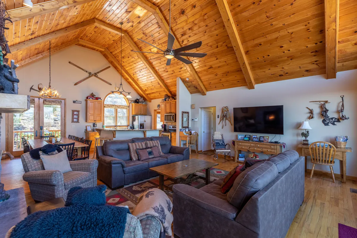 A wide view of the grand gathering space featuring a vaulted wooden ceiling, a large sectional sofa arranged around a prominent stone fireplace, and an adjacent dining area near the open kitchen. The warm wood finishes, ample seating, and central fireplace capture the heart of communal living and relaxation.