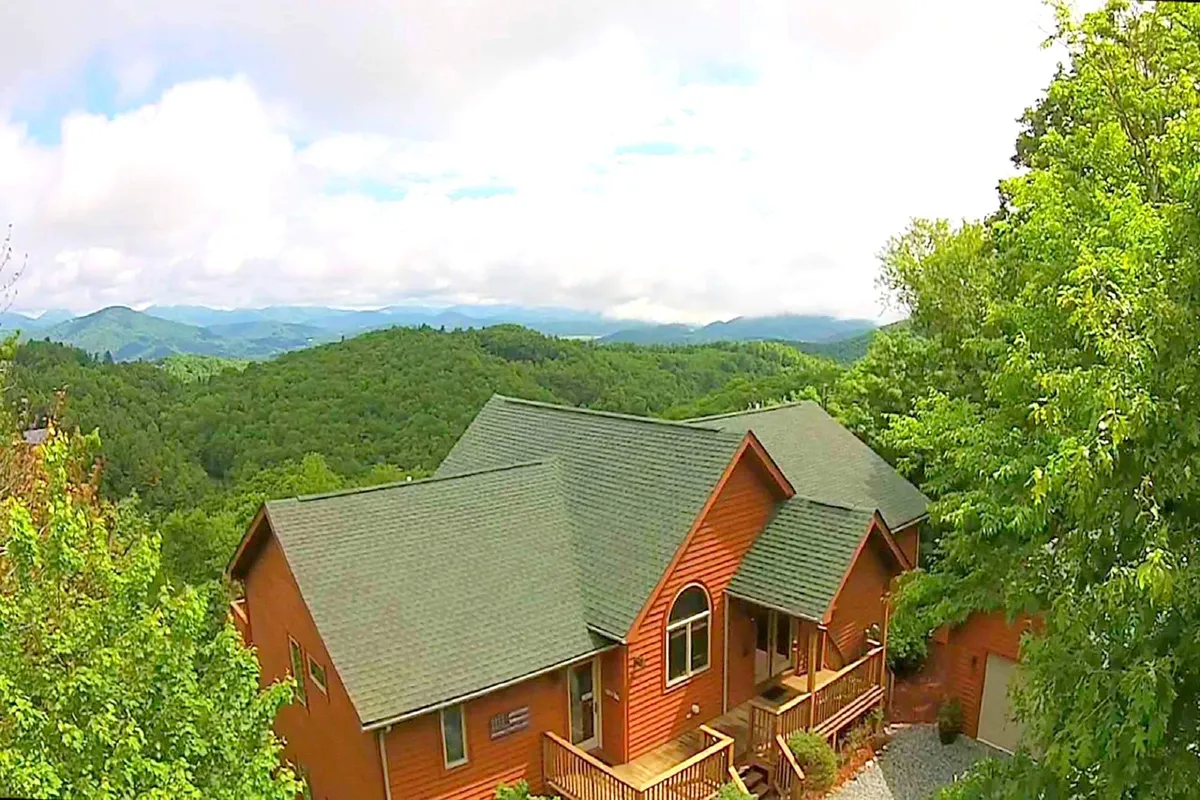 The cabin's wooden exterior is set against green mountain slopes and features a deck area with sturdy railings. The setting conveys tranquility and the sense of being immersed in nature, with room for both people and pets to enjoy the outdoors safely. The mountains in the background hint at nearby trails and adventure.