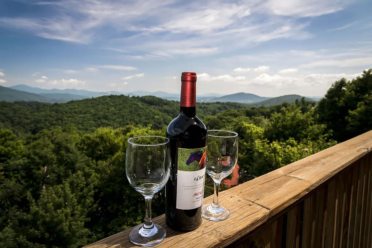 A close-up of a wine bottle and two glasses set on a balcony railing, with a dramatic mountain vista in the background. The scene evokes a sense of intimate relaxation and indulgence.