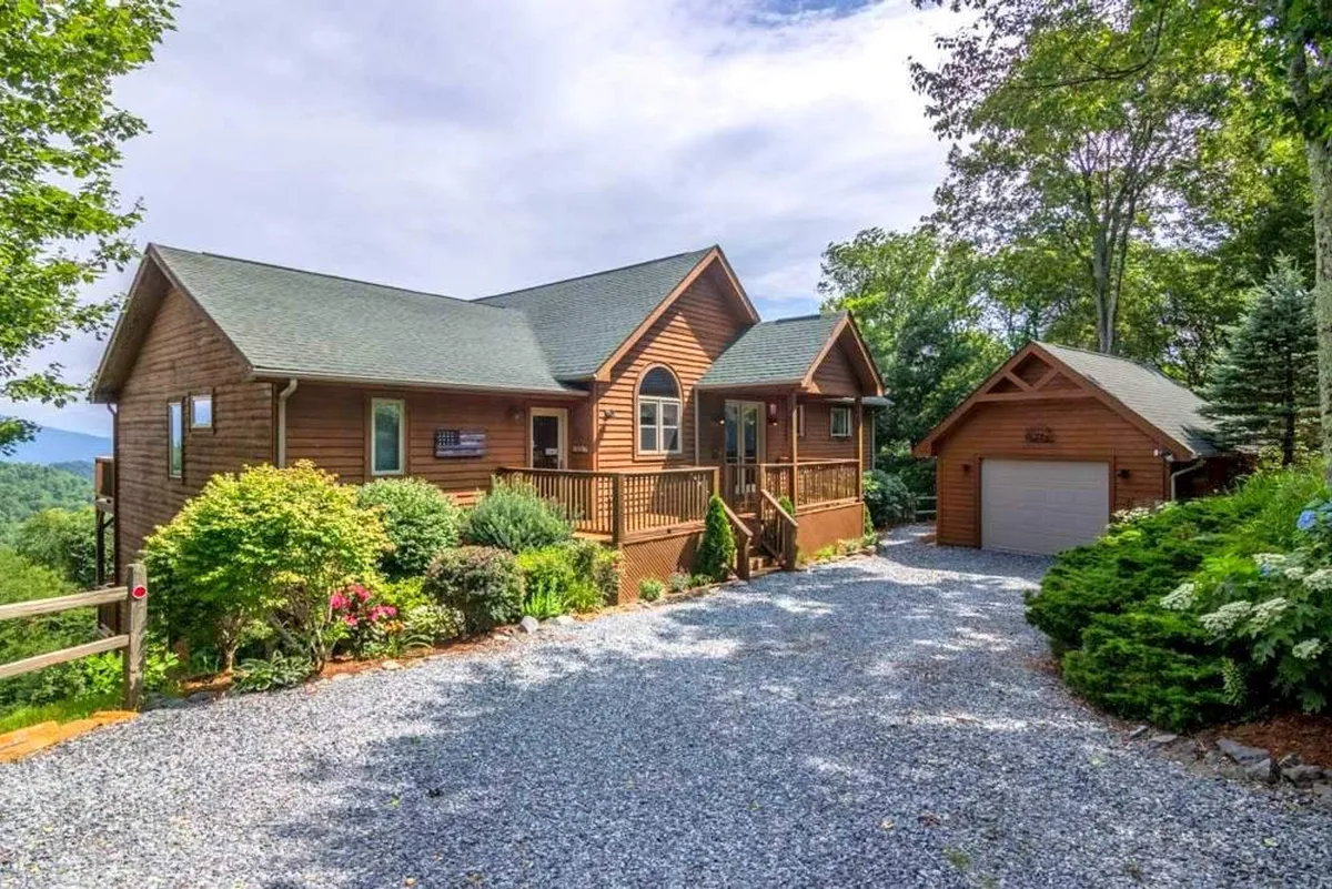 The image showcases the cabin's inviting wooden facade framed by lush greenery and colorful flowers, a spacious gravel driveway leading up to the house, and an attached garage. The front porch offers ample space for relaxing while taking in the surrounding nature.