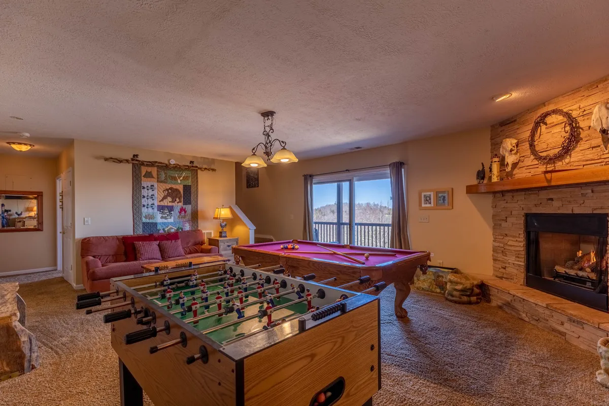 A spacious game room featuring a foosball table and a pool table, with a stone fireplace providing a cozy backdrop. A large window lets in natural light and offers a view outdoors, enhancing the vibrant and playful mood. The arrangement of the tables suggests ample space for group play and social gatherings.