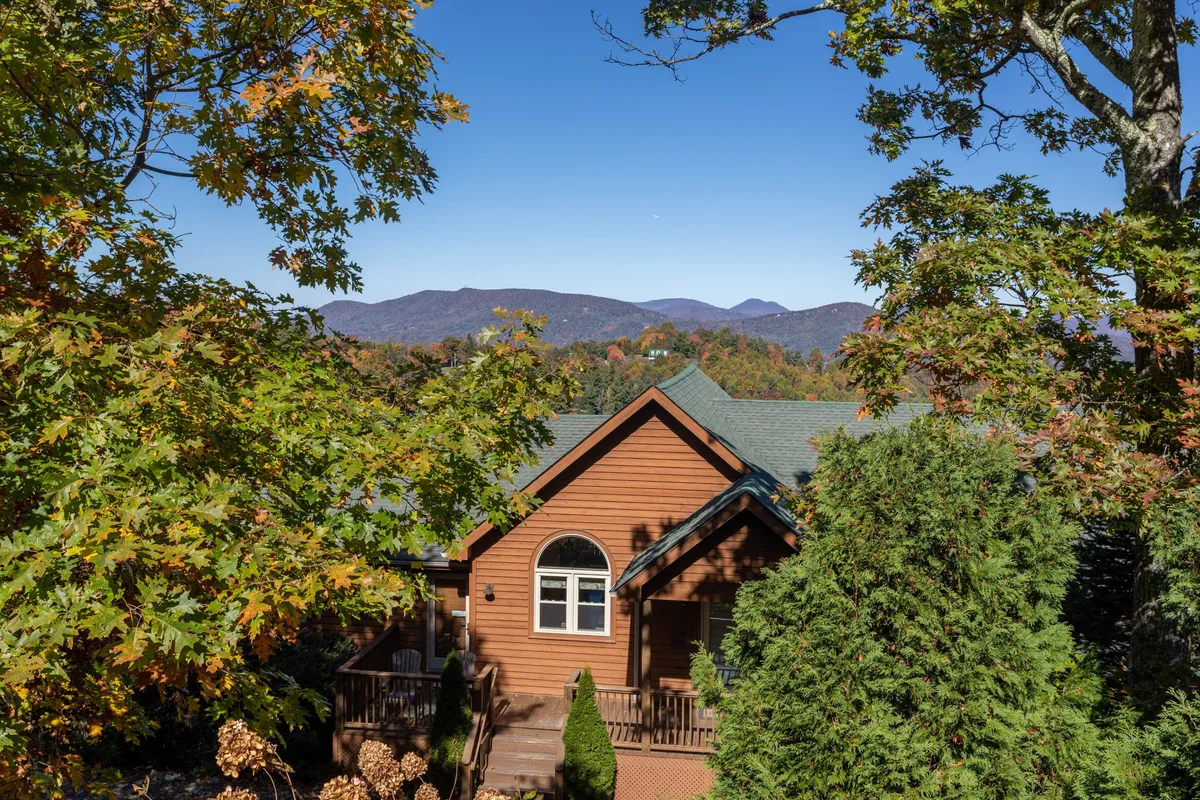 This image features a welcoming wooden house surrounded by lush trees, with a dramatic mountain backdrop. The setting captures the sense of being nestled in nature, while the positioning of the house and visible porch suggest immediate access to outdoor adventures and trails.
