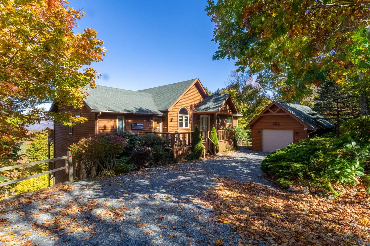 A charming wooden house sits nestled among lush greenery, with a vibrant garden and colorful autumn leaves surrounding the property. A driveway leads up to the welcoming porch, suggesting easy access for guests and their pets. The outdoor space appears expansive and inviting, with plenty of room for enjoying nature.