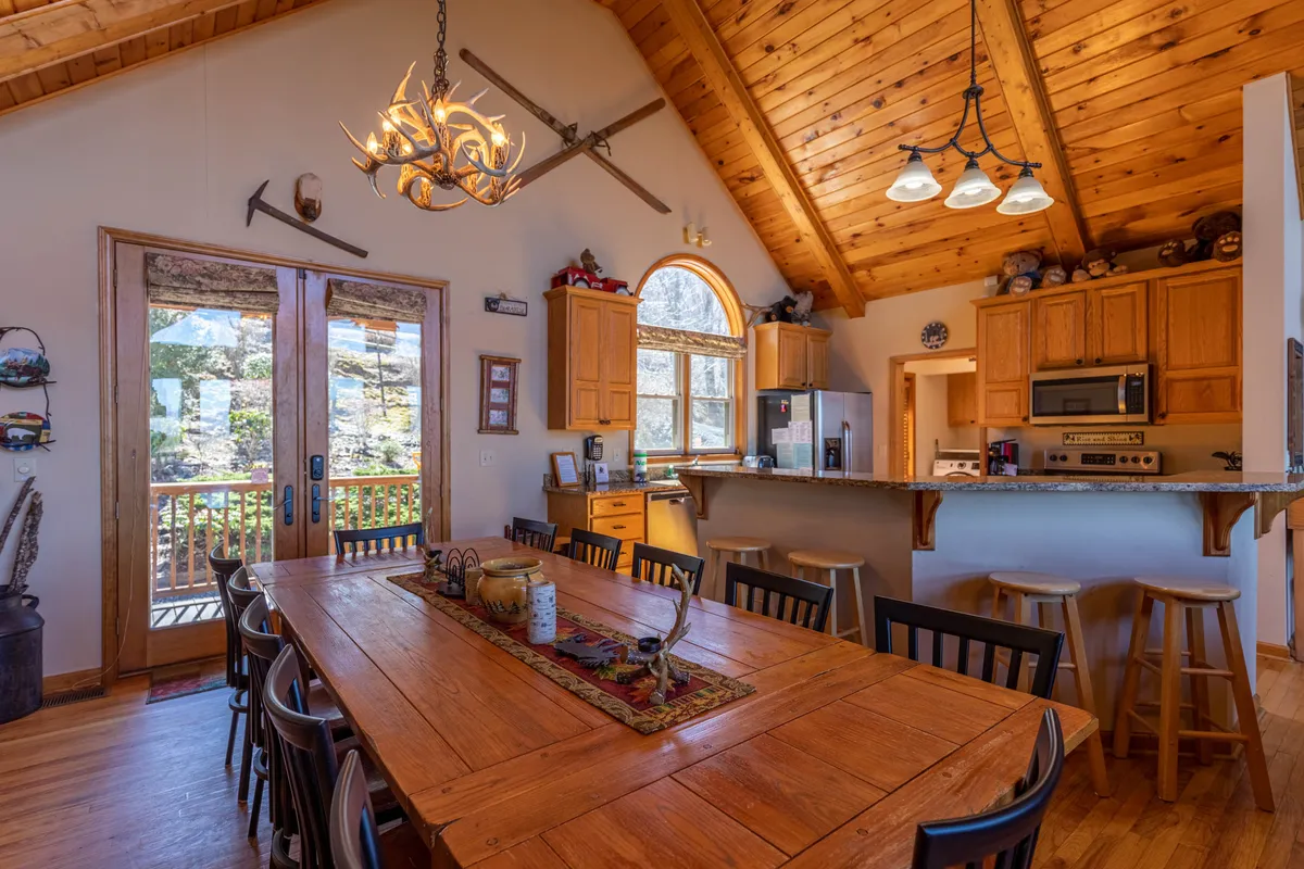 A bright and welcoming dining area with a large wooden table that comfortably seats twelve, illuminated by a chandelier and natural light from spacious windows. The open transition to the kitchen highlights bar seating and granite countertops, underscoring spaces designed for shared meals and celebrations.
